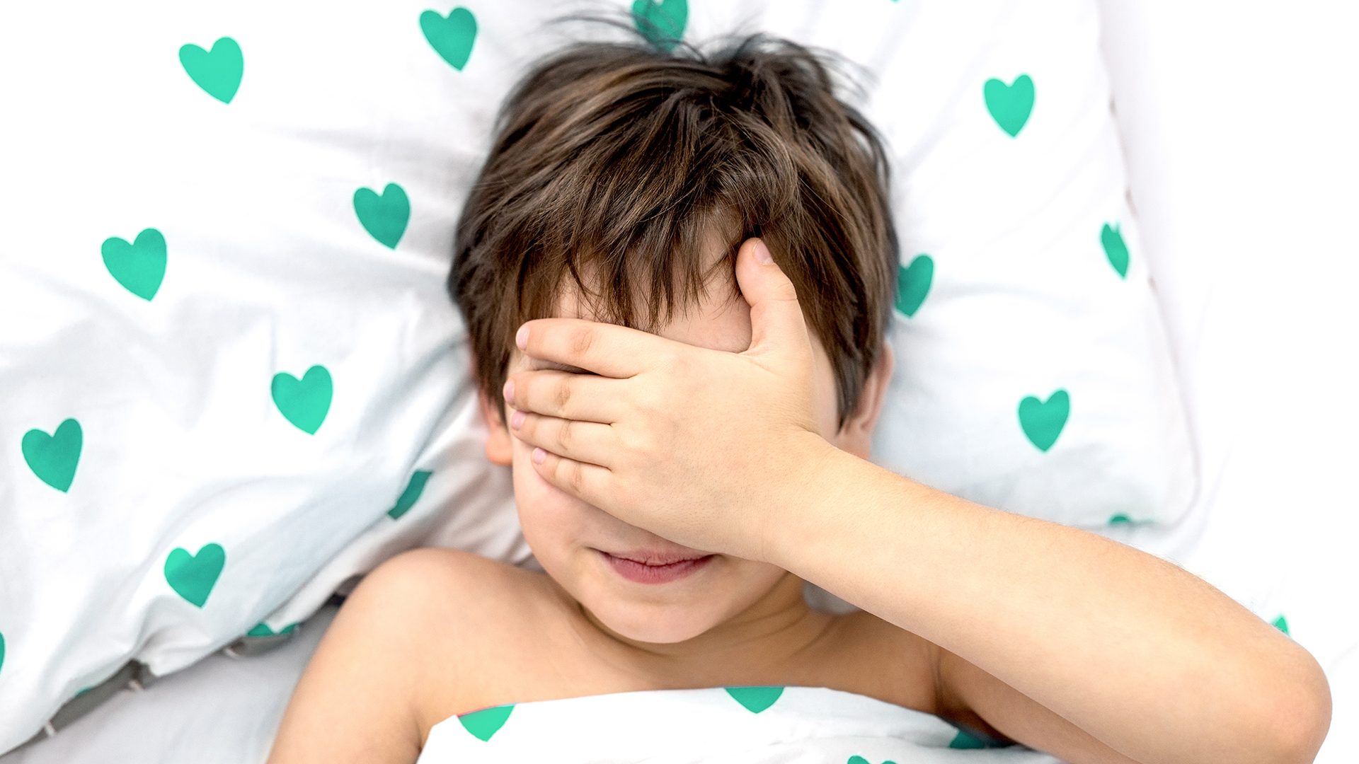 the boy lies in a bed with green hearts, his face in his hands. emotions without a face. white color, top view